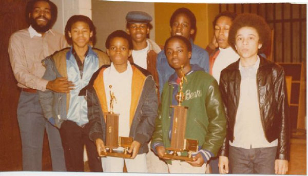 Color photograph of an adult and group of young, African American boys surrounding two young men holding basketball trophies, one adult stands