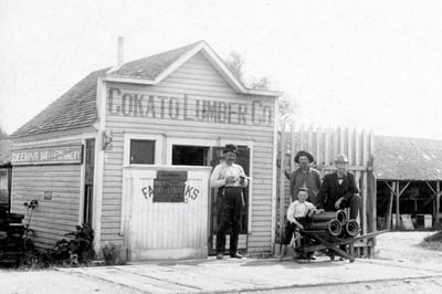 Building with a sign saying Cokato Lumber Co. with a man standing in front of it, next to him are two men and and boy staining around a wheel barrow loaded with clay pipes
