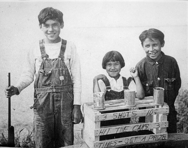 Two Ojibwe boys and a girl standing by crate with writing 3 SHOTS 10 Cents HIT THE CANS 3 TIMES - GET A PRIZE. Oldest boy is holding a rifle