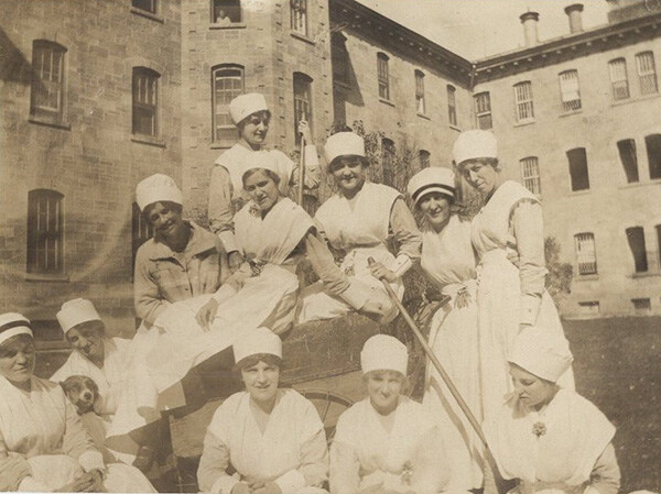 Group of women in nurse uniforms sitting on and around wagon in front of hospital building