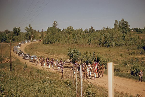 _Color photo of the 1955 Memorial Day in Sandstone, MN-Brushland and dirt road with honor guard leading marching band and long row of cars copy