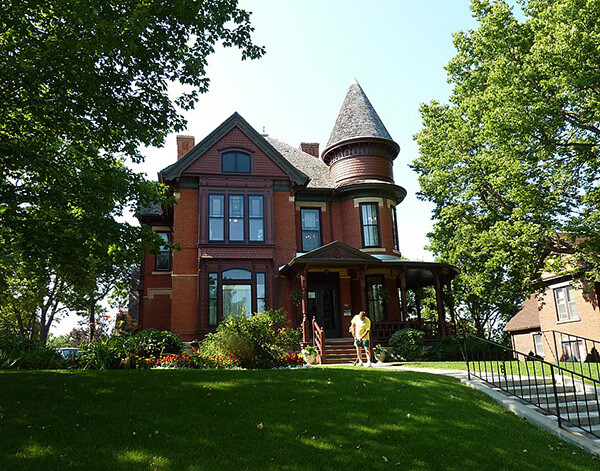 Exterior of the John Lind House.  A man is trimming the grass along the pathway to the house