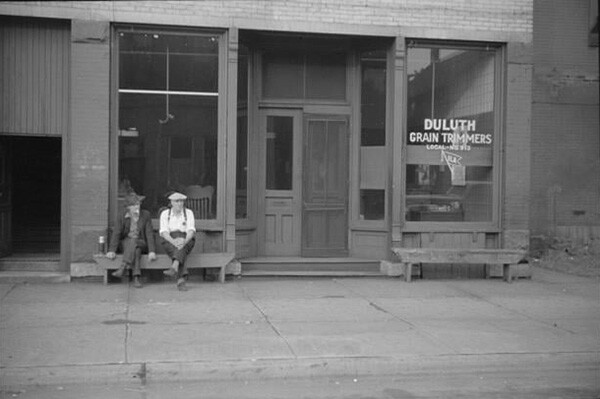 Storefront with window sign, DULUTH GRAIN TRIMMERS. Two men are sitting on bench in front of store