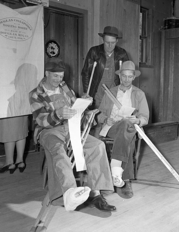 Three farmers on crutches looking at ballots. Voting booth in background