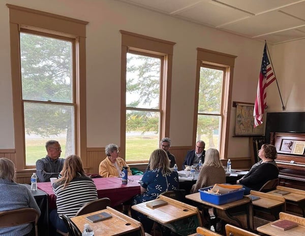 nine people sitting around a table in an old school house room having a meeting