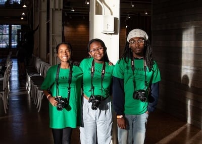 Three young African Americans holding cameras and wearing green MAAHMG t-shirts