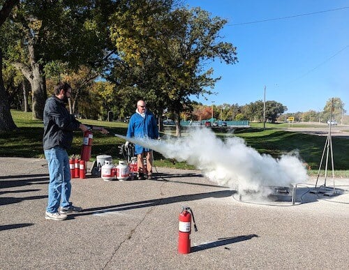 In a workshop, a person using a fire extinguisher to put out fire with another person looking on