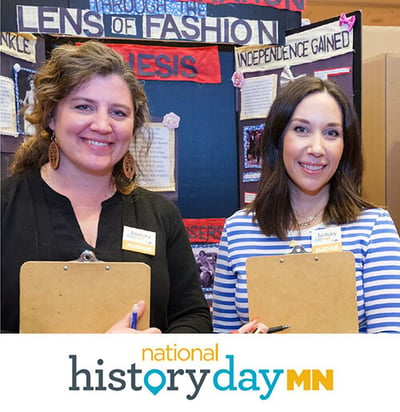 Two Judges standing with clipboards in front of history displays with MN History Day Logo under photo