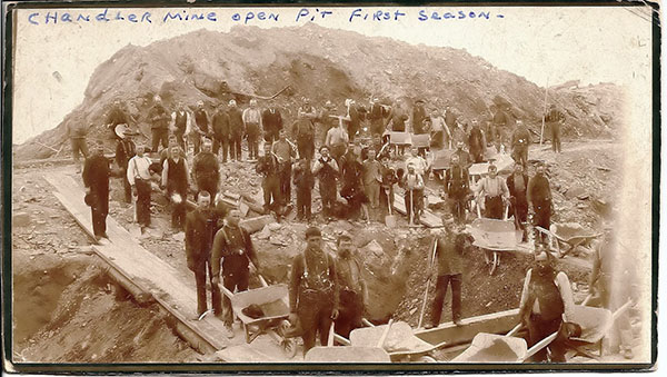 Photo of men with wheel barrels in a pit with a large mountian of dirt behind them. On the photo is written, Chandler Mine open pit first season