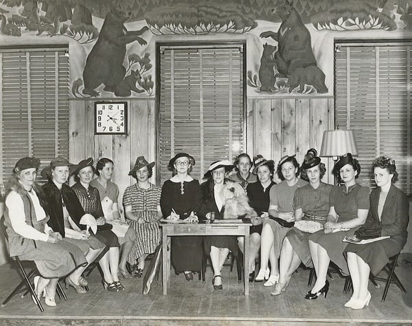 Photo is of 12 women sitting around a small writing desk. This is the cast of the Milaca Civic Clubs play-Meeting to Music