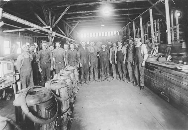 Nearly 20 men in workclothes standing in workshop foundry, in front of them are a row of nearly completed cast iron stove