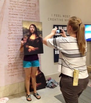Photo of a woman taking a photo of another woman who is holding up a posterboard of the Mona Lisa painting with her face sticking out in a cut out hole where the Mona Lisa's  face should be.
