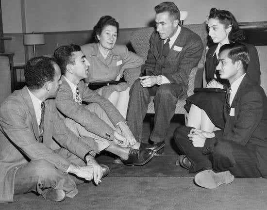 Title: Thanksgiving Feast for University's Muslim Students. Black and White photo of a group of four men in suits and two women in dresses sitting in a semicircle. Three are sitting on floor, one man is sitting in winged back chair. ca.1940s.