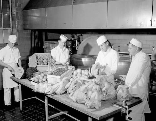 Black and white photo of four men in white cook uniforms standing in a commercial kithen  prepping a table of turkeys and other food stuffs.