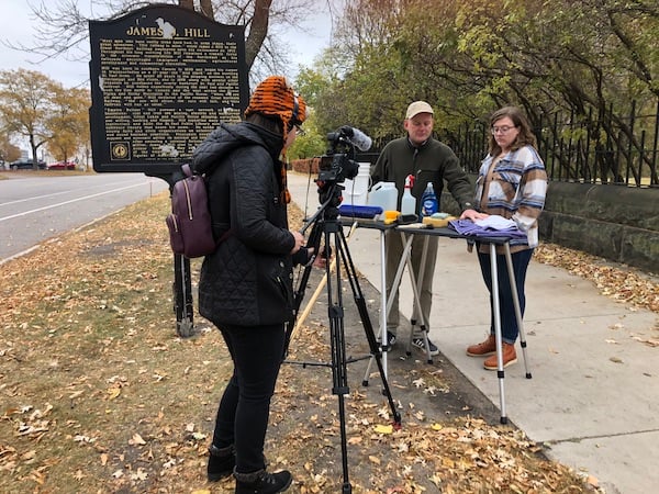Woman standing on boulevard holding video camera with camera tripod in front of her.  Next to her is black cast aluminum historical marker. Near it is a table holding cleaning supplies. Two people are behind the table.