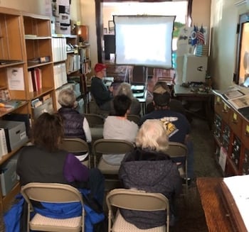 Nine people sitting in small room tightly packed together watching presentation screen.