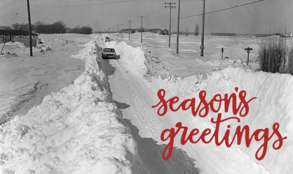 Car traveling down long and narrow road surround by very high snow drifts-Seasons Greettings is superimposed on top of photo