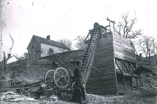 ca1900 photograph showing outdoor operation of a converbelt machine. Appears to be moving material (gravel?) from a lower level to a top of a two-story wood inclosed structure on the upper level. Their appears to be three chutes on box