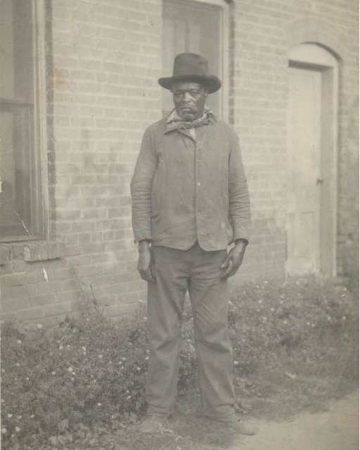 African American man wearin hat and a handkerchief around neck, standing in front of brick building with a serious look on his face. ca. 1890s