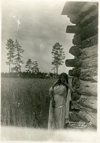 Black and White photo of young woman in white smock dress leaning against log cabin. Wheat field in the background with pine trees in far distance copy