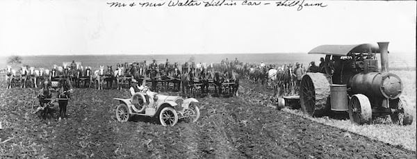 White Packard automobile in middle of plowed farm field behind them are roughly two dozen teams of horses. To the right of car is a steampowered tractor-ca. 1908