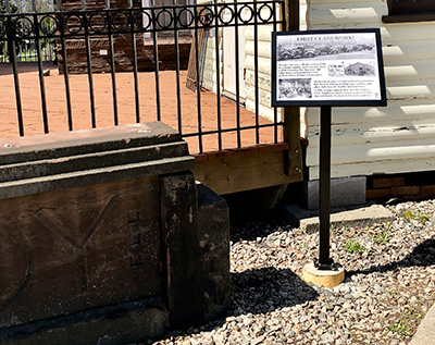 To the left of the signage is a large old sandstone sign from a building placed on the ground. Behind the signage is a wooden building