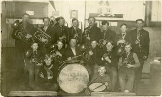 Black and white photo of the Prinsburg Band standing for portrait. 16 members with insturments wearing suits and standing in what appears to be the room a one-room school house