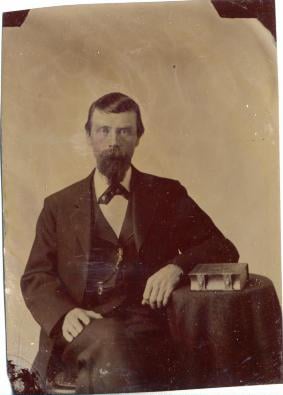 Tintype of a man looking at the camera, sitting with is arm resting on a small table that has a book on it.