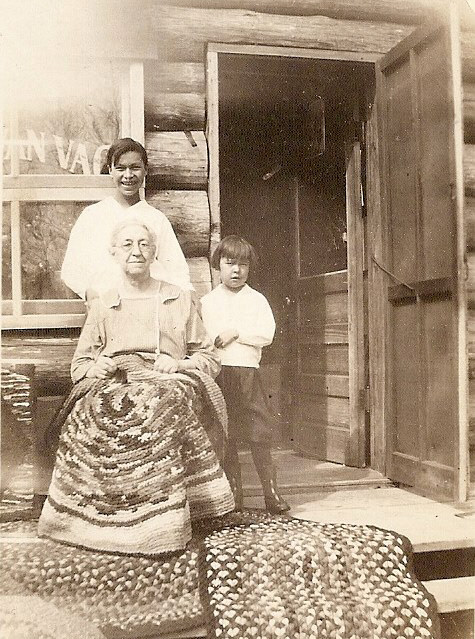 Two Native American children and old woman. Woman has rug on her lap and two rugs on the ground in front of her. They are standing outside a log building