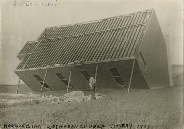 1908 photo of a church tipped upside-down with a man standing in front of it. Text on the photo says, Norwegian Lutheran Church Dovray 1908.  Built 1898