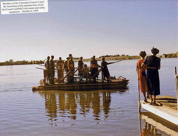 Women on dock watching pontoon boat with 10people on it. Sticker on photo reads Members of the Volunteers Council watch the launching of the pontoon boat which the Council outfitted with motor and safty equipment - October 6, 1959.