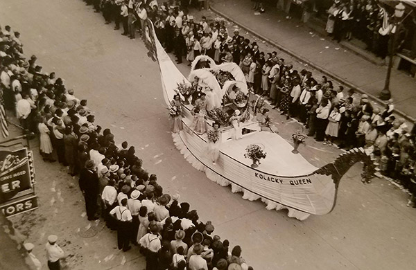 1940 Black and white photo. View looking down at a parade float in the shape of a white Viking Ship. On the bow of the ship it says Kolacky Queen. Queen candidates sit on the float and wave at the crowds