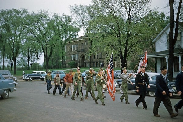 ca. 1950 color photo of a Parade with veterans in front and Boy Scouts following carrying an American flag in the background is a school building