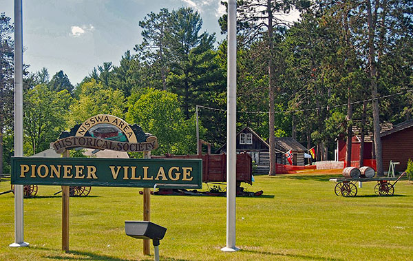 Grounds with large sign saying Nisswa Area Historical Society Pioneer Village in foreground with historic buildings and wagons in the background