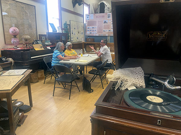 in background three people discussing around a table in a museum gallery, foreground is a victrola