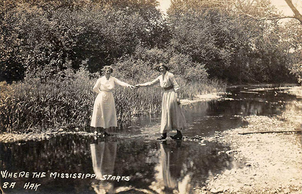 Photo of two women in victorian dress holding hands over a stream. Text on photo says Where the Mississippi starts