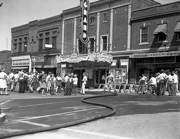 A Crowd of children stand outside on the street in front of the Grand Theater. Adults are to the left of them and a fire hose is to the right going into the theater.