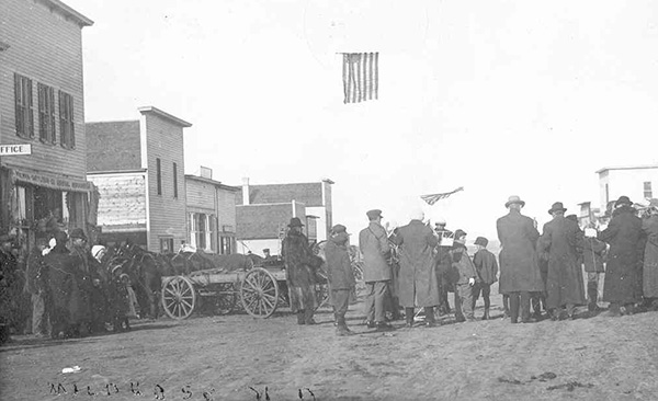A small town main street in early 1900s with a crowd of people standing in the middle in winter coats playing musical insturments - flags fly above them