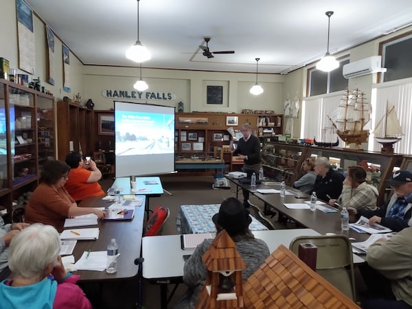 Meeting inside a museum gallery. Man speaking at the front of audience with powerpoint presentation. Audience sitting at tables.