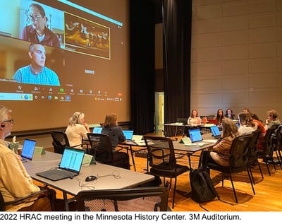 HRAC committee meeting on auditorium stage. In background is a large video screen with members that are meeting online-text