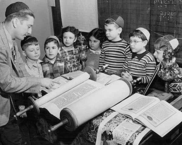 Rabbi talking to a group of young students while pointing to a Torah and other books on a table