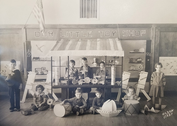 1937 photo of school classroom with eight 3rd grade students standing in a handmade toyshop with cardboard toys. Above is a sign saying Our Little Toy Shop