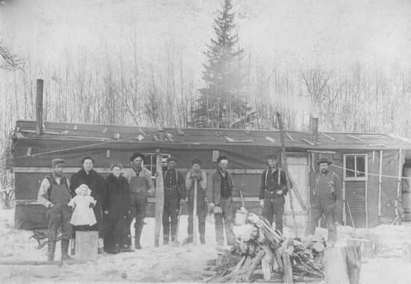 Winter photo of the Blakely Logging Camp. & men and 2 women and a infant standing infront of a long shack. A small pile of wood is in front of them. ca 1900