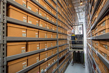 Corridor showing rows of shelving filled with boxes. A worker on a lift is retrieving a box from a shelf