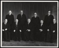 Nine US Supreme Court Justices sit and stand in two rows. They wear black robes.