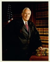 A white-haired man stands next to a desk with three books on it. He wears the robe of a Supreme Court justice. Behind him area an American flag and a bookshelf.