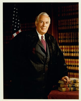 A white-haired man stands next to a desk with three books on it. He wears the robe of a Supreme Court justice. Behind him area an American flag and a bookshelf.