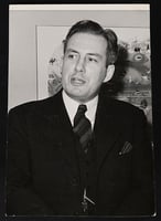 A standing man wearing a suit and tie, shown from the waist up to his head. Behind him is a piece of art hanging on a wall. 