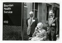 Ruth Boynton with colleagues Don Cowan and Paul Rupprecht at the ceremony where the University Health Service was renamed the Boynton Health Service. She is elderly and sitting in a wheelchair. The two men stand behind her and the sign with the name of the building is in the foreground.