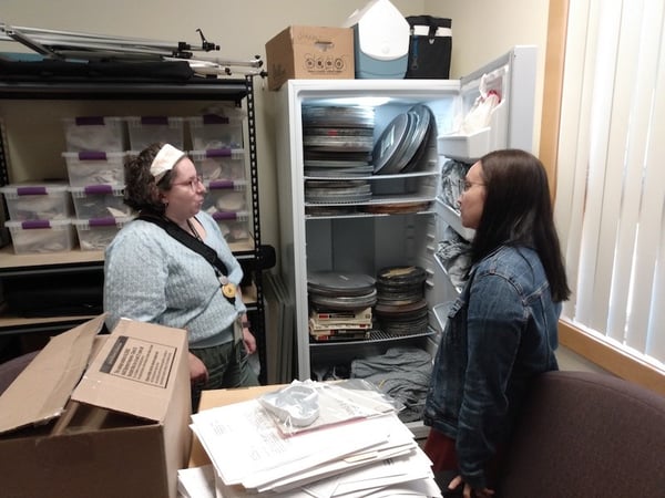 Two people standing talking in collections storage room next to open freezer with film canisters inside